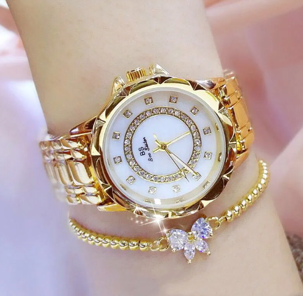 Women's elegant gold color watch with white table and sparkling details shown with jewelry next to it
