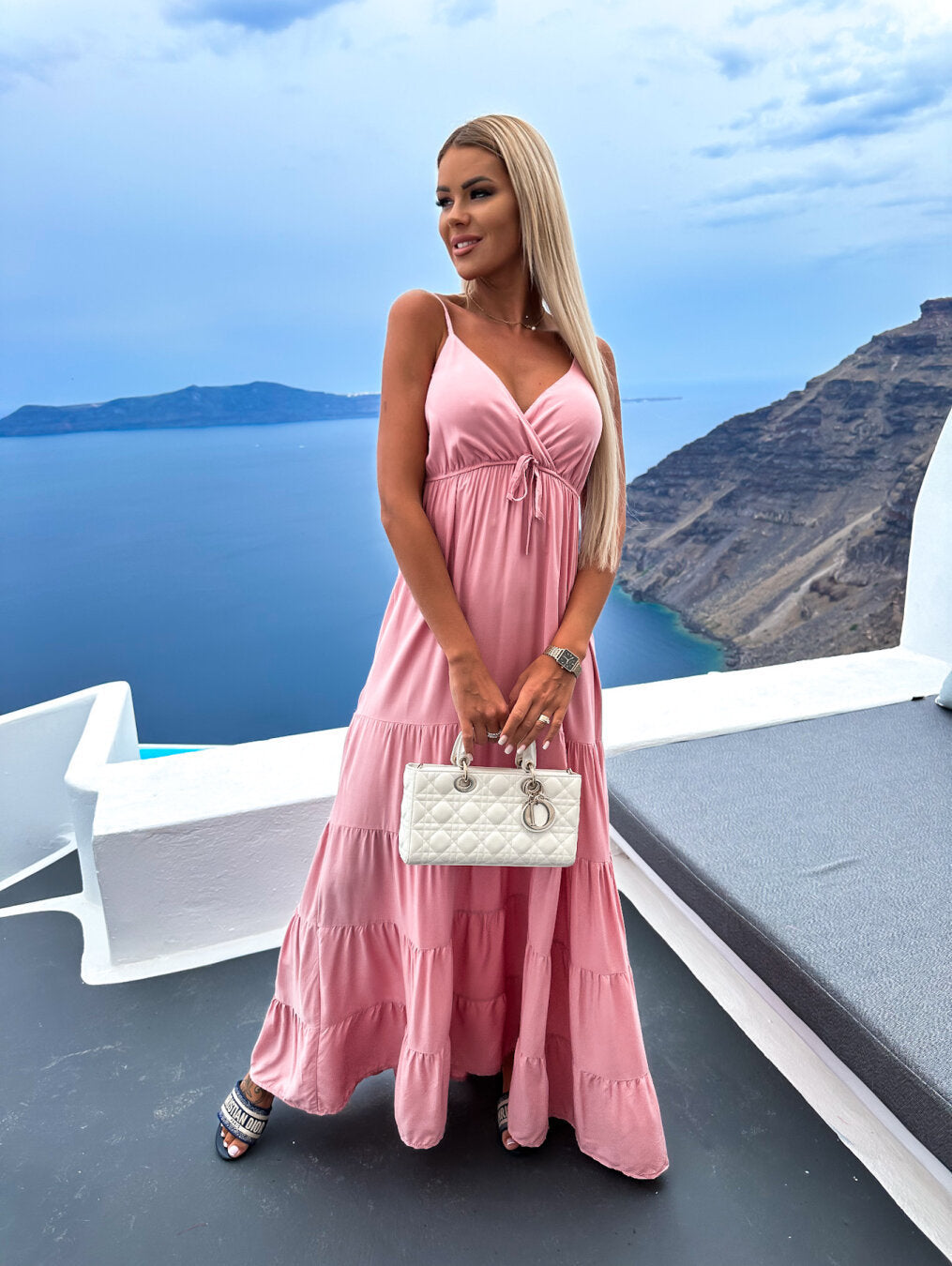 Model wearing pastel pink ruffle-hem maxi dress with white handbag, feminine vacation outfit
